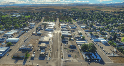 Aerial view of a small town with a main street lined by buildings, surrounded by green fields and distant hills.